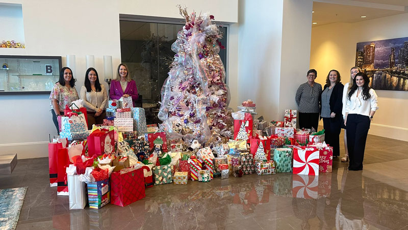 Seven adults posing with a Christmas tree surrounded by wrapped holiday gifts in an office setting